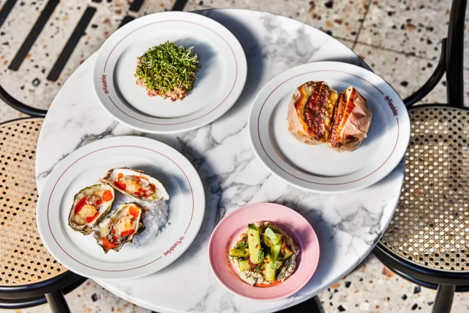 Overhead shot of plates with oysters, vegetables, potato, and sprouts on a white marble table, casting soft shadows from natural light.