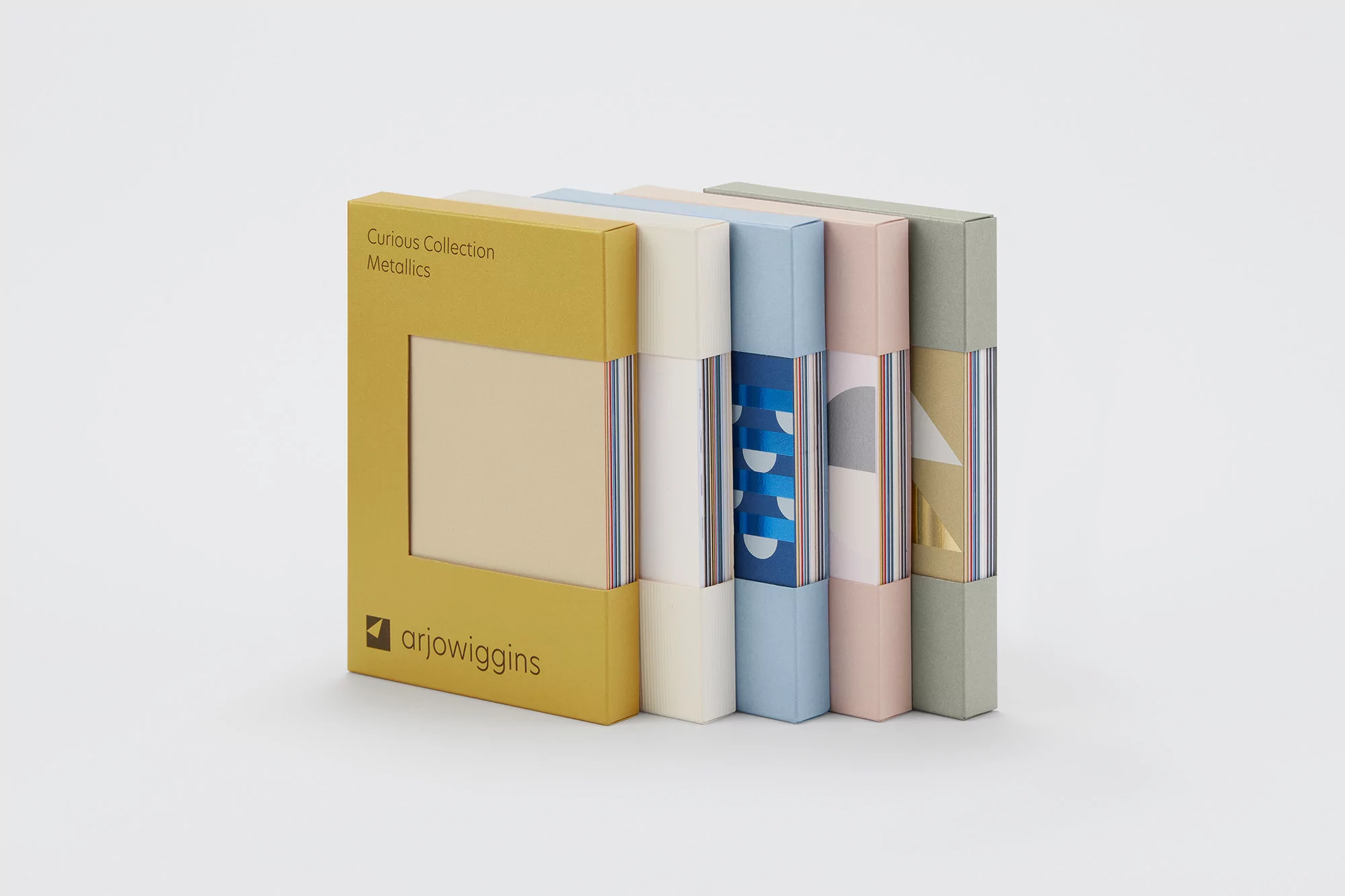 Stacked boxes of Curious Collection Metallics paper samples in mustard yellow, cream, blue, light pink, and sage on a white background.