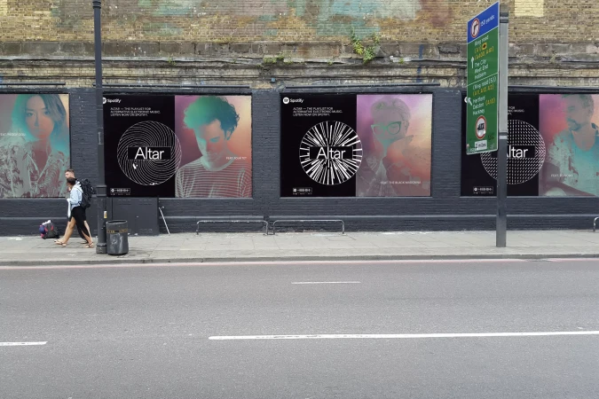 Outdoor posters on a brick wall feature Spotify's Altar playlist with faces. Pedestrians walk along the sidewalk near a city street.