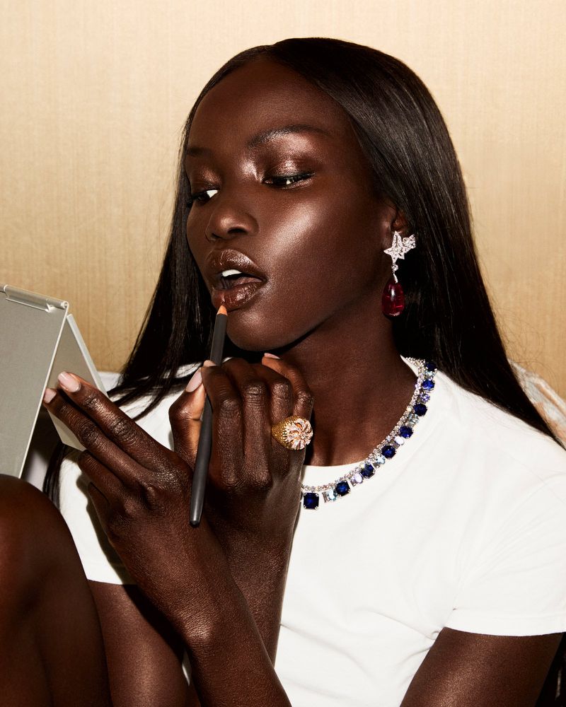 Elegant dark-skinned woman applies lip liner in studio, showcasing jewelry, beauty, and fashion in a close-up portrait-style shot.