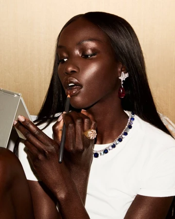 Elegant dark-skinned woman applies lip liner in studio, showcasing jewelry, beauty, and fashion in a close-up portrait-style shot.