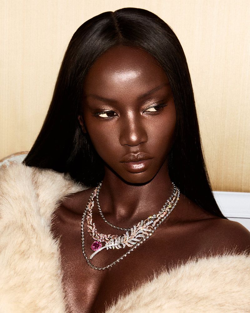 Close-up shot of a woman with dark skin wearing layered necklaces, posed against a neutral backdrop with soft lighting and fur stole.