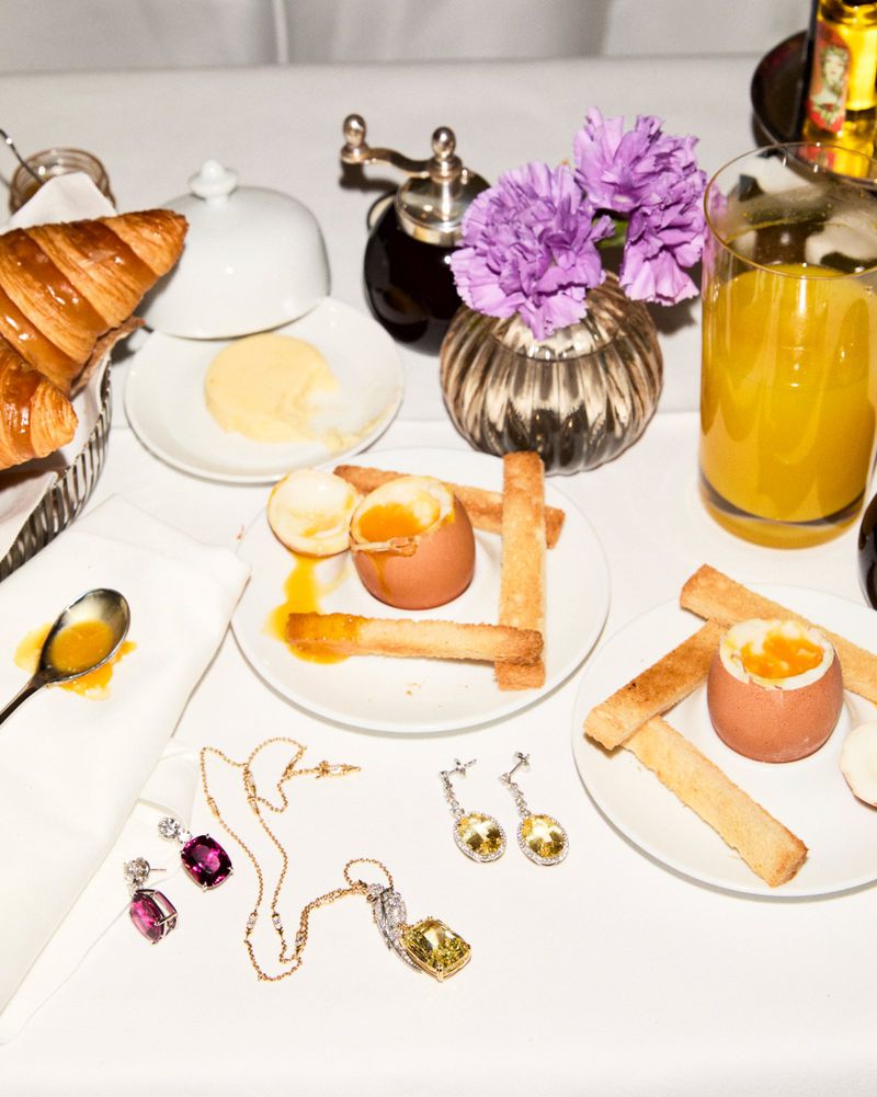 Breakfast still life featuring croissants, soft boiled eggs, orange juice, and luxury jewelry on a white tablecloth.