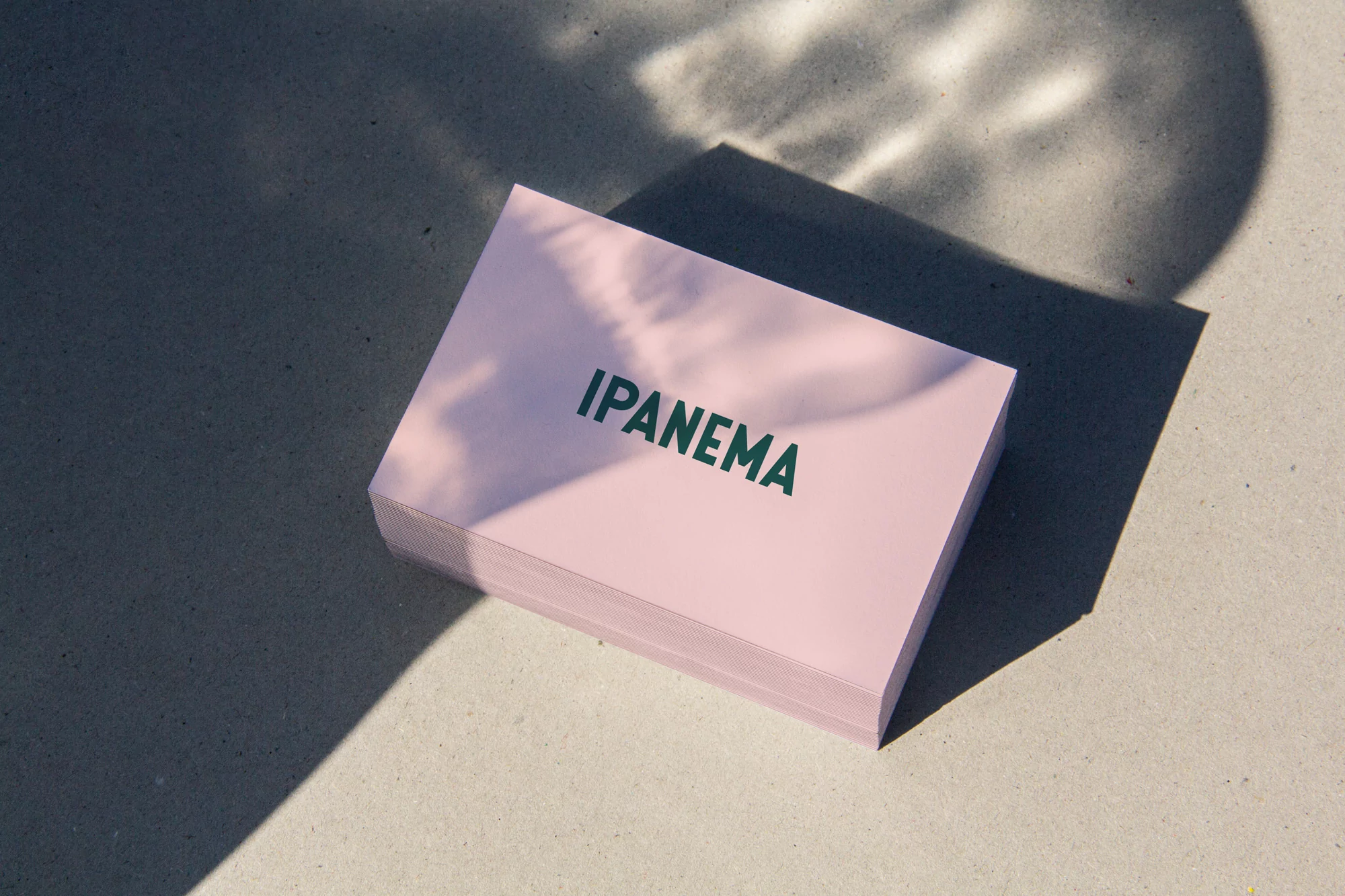 Stack of pink paper sheets with 'Ipanema' text, casting shadows on a grey surface, creating a minimalist, graphic design aesthetic.