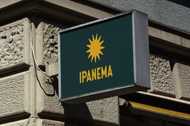 Hanging rectangular storefront sign for Ipanema with a yellow sunburst logo against a textured stone building exterior