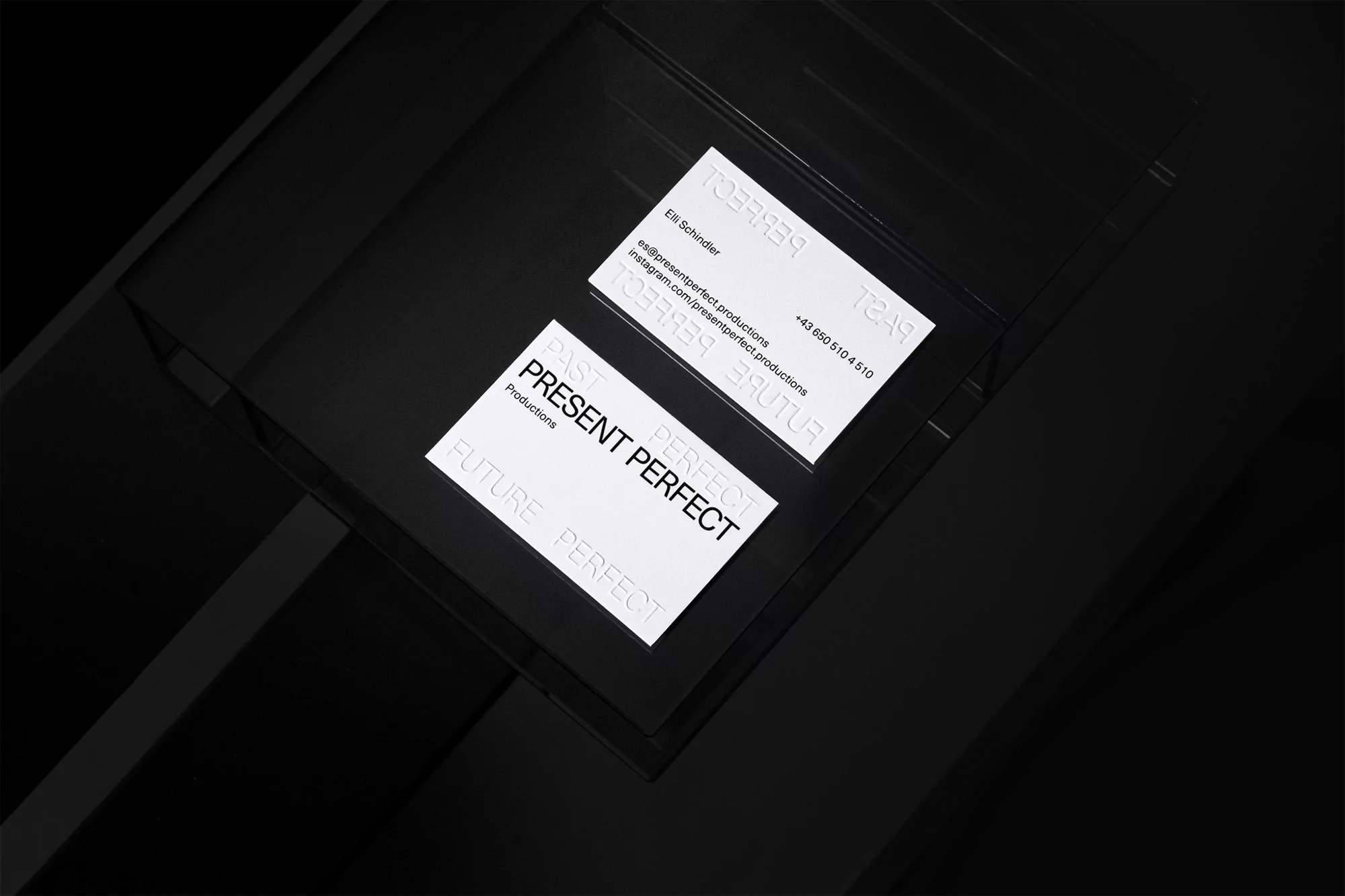 Two business cards sit on a black surface with 'Present Perfect Productions' visible. Minimalist design, studio photography, high-contrast.