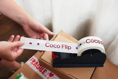 Person unrolling branded packing tape with red text from a dispenser onto a cardboard box