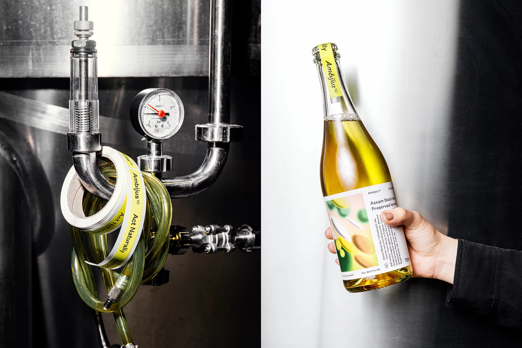Close-up studio shot of a hand holding a bottle of Ambitus artisanal beverage, set against stainless steel equipment and industrial pipes.