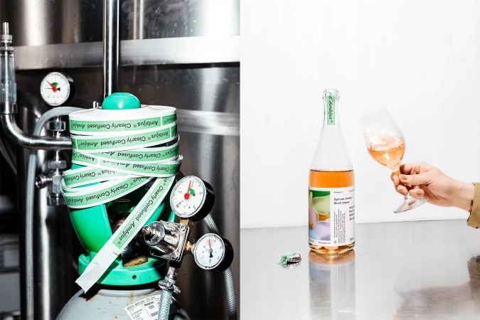 Bottle of artisanal fruit wine on reflective surface with hand holding glass, next to brewing equipment wrapped with branded tape.