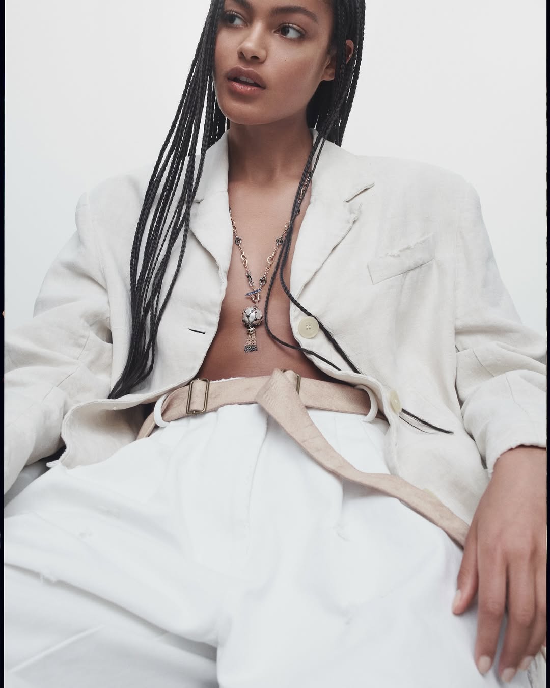 Young woman with braids in a blazer and white pants, adorned with a necklace and light-colored belt, posing in a neutral setting.