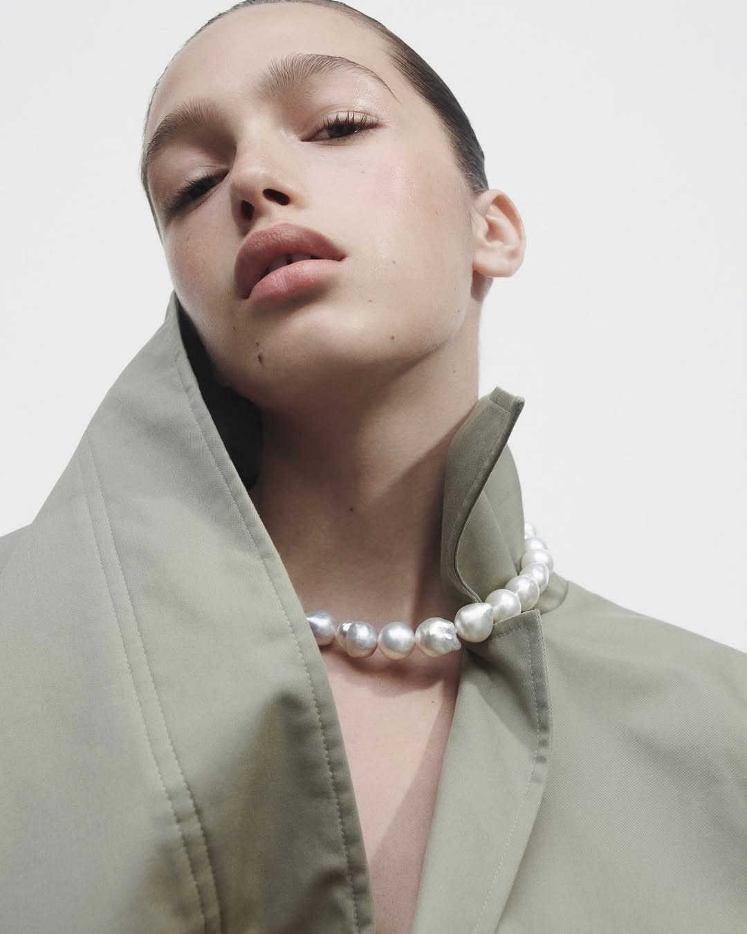 Close-up of a young adult woman with a pearl necklace and light green jacket against a bright background, showcasing beauty and fashion.