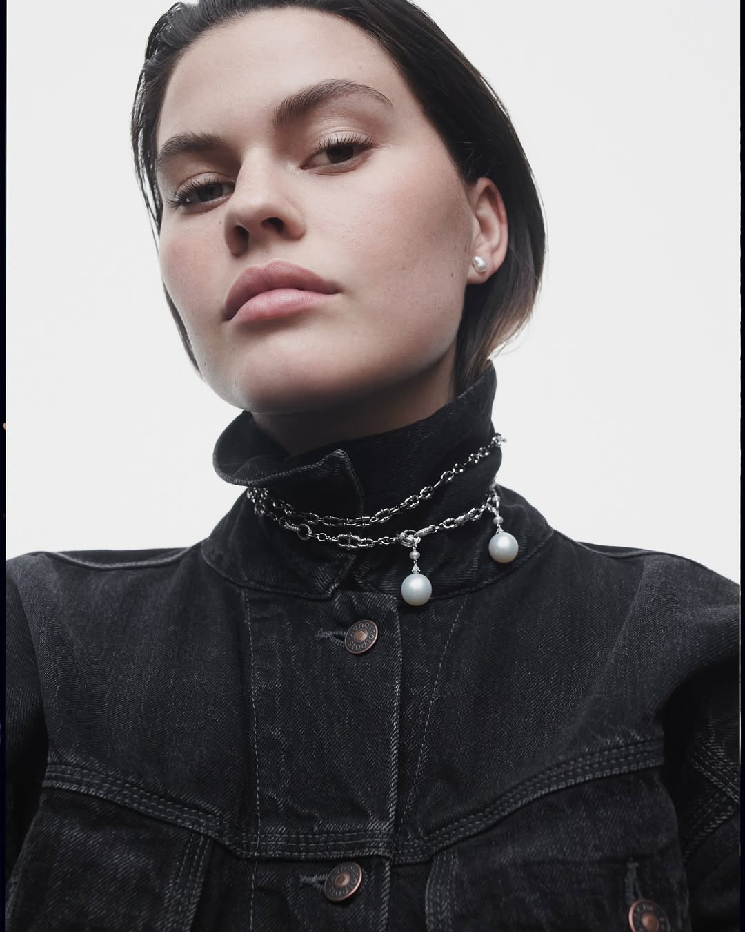 Low-angle portrait of a woman in a black denim jacket wearing silver chain necklaces with pearl pendants against a light background.