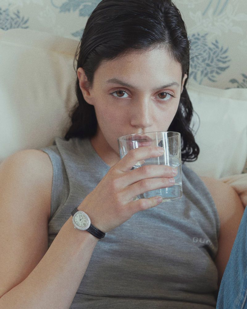 Young adult woman in a grey tank top sips from a clear glass, wrist watch visible, indoors, soft lighting, floral wallpaper backdrop.