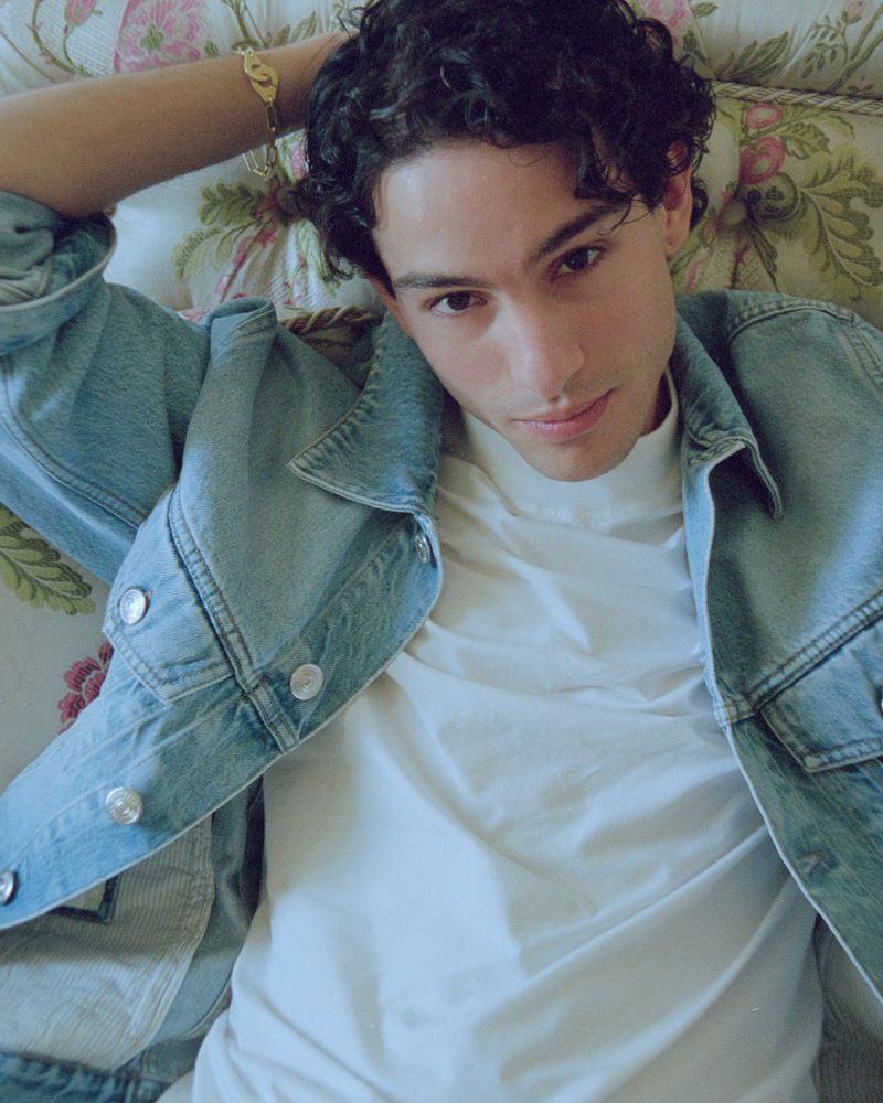Young adult male poses in a denim jacket and white t-shirt against a floral background, lying down and looking at the camera.