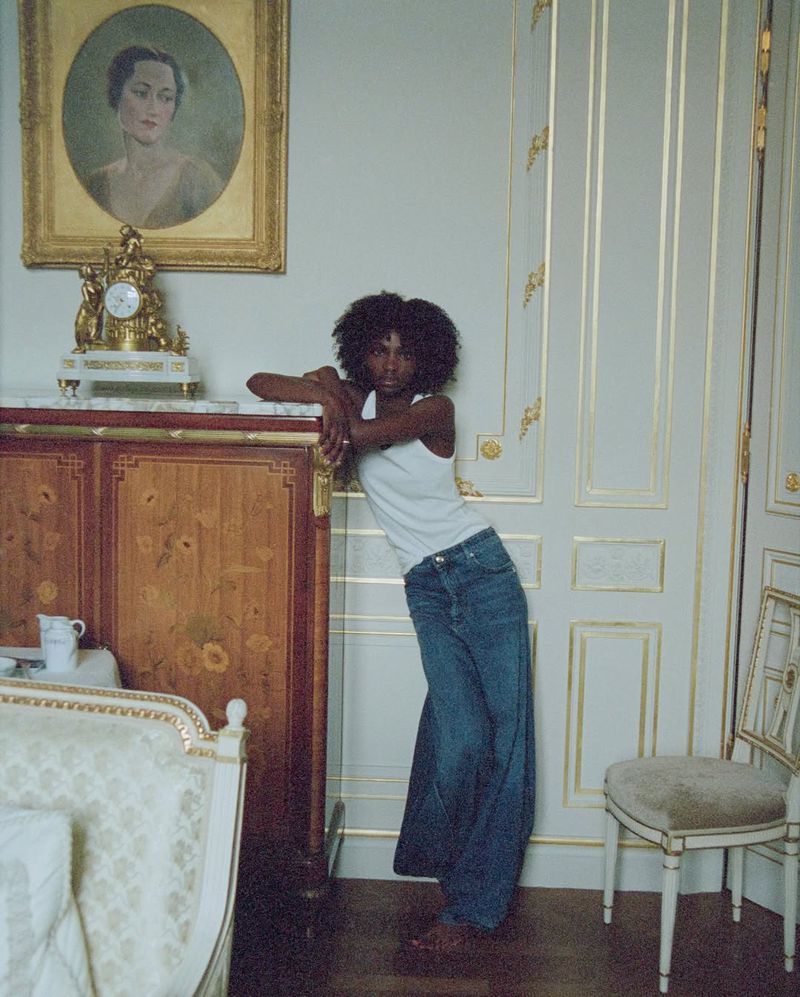 Young Black woman in a tank top and jeans leaning against wooden cabinet in a luxury home interior with a portrait in the background.