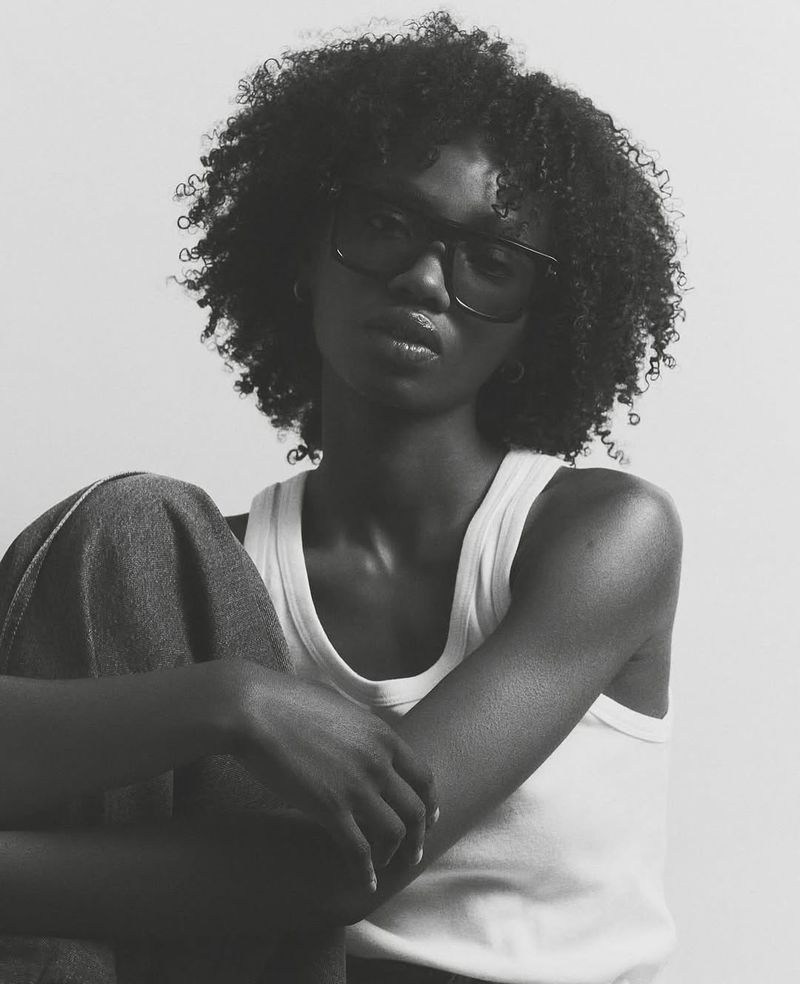 Monochromatic portrait of a Black woman with curly hair, wearing a white tank top and glasses, posing in a light-colored indoor setting.