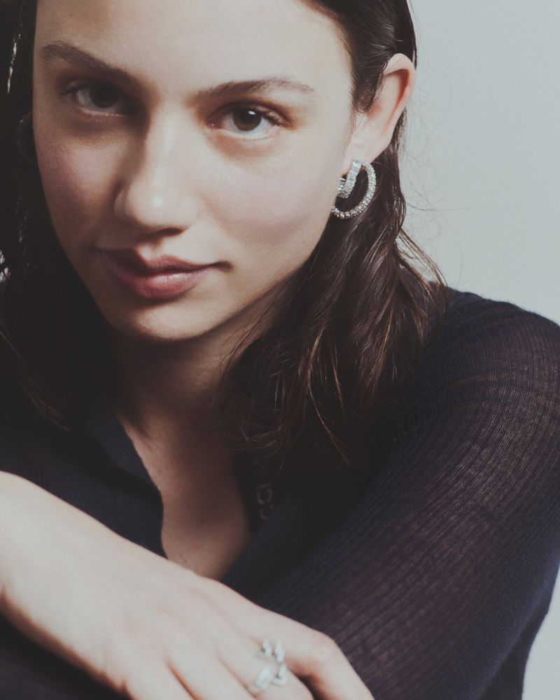 Close-up of a young woman with dark hair, wearing earrings, and a dark top. The image features soft lighting and a neutral background.