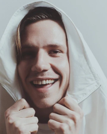Close-up studio shot of a smiling young adult male wearing a white hooded jacket, showcasing his teeth and casual fashion.