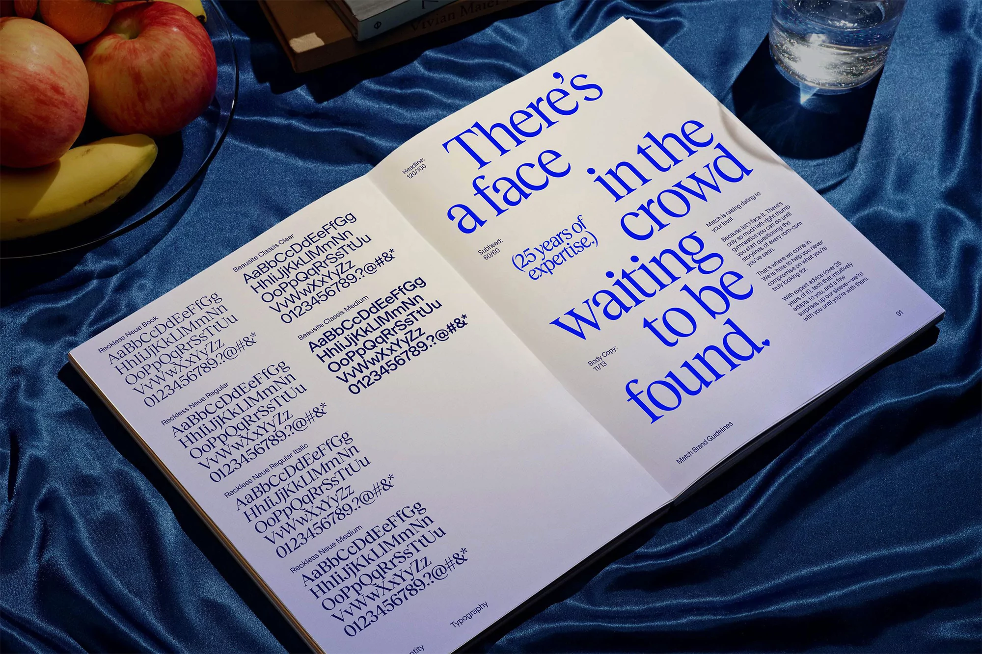 Open book on a blue fabric surface displays typography with bold blue fonts, bananas, apples, and a glass of water in the background.