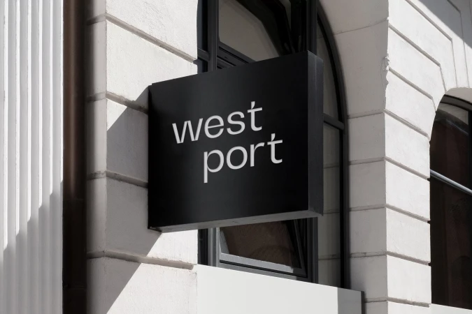 Exterior shot of a black sign with white text that reads 'west port' hanging on a white stone building with black window frames.