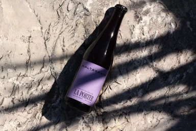 Dark glass beer bottle with a purple label resting on a rough grey stone surface outdoors in harsh sunlight