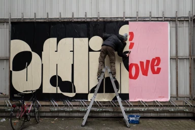 A person standing on a ladder pasting large black and cream typography posters onto an outdoor wall.
