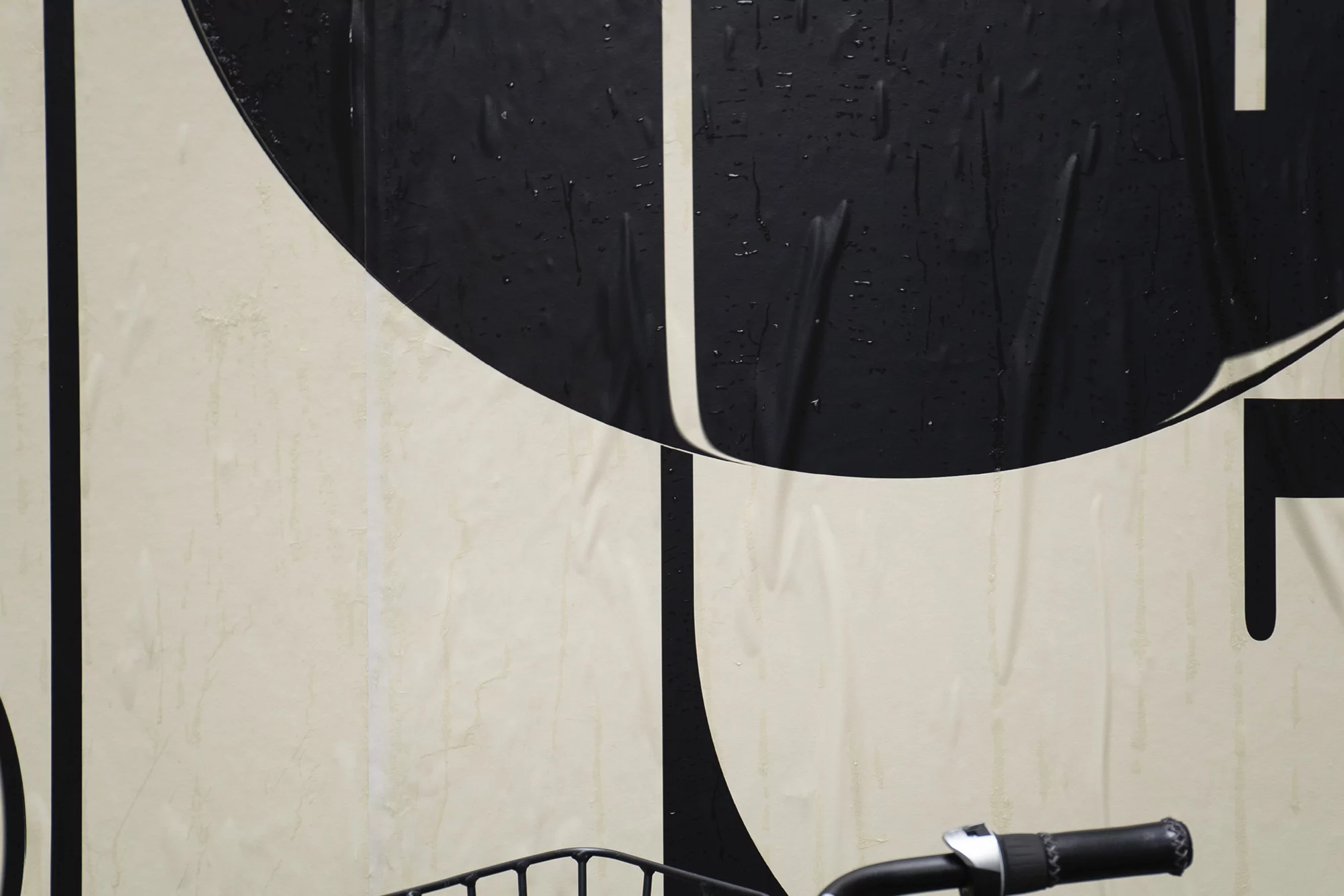Close-up of a bicycle handlebar and front wire basket against a damp poster-covered wall with geometric black and cream shapes.