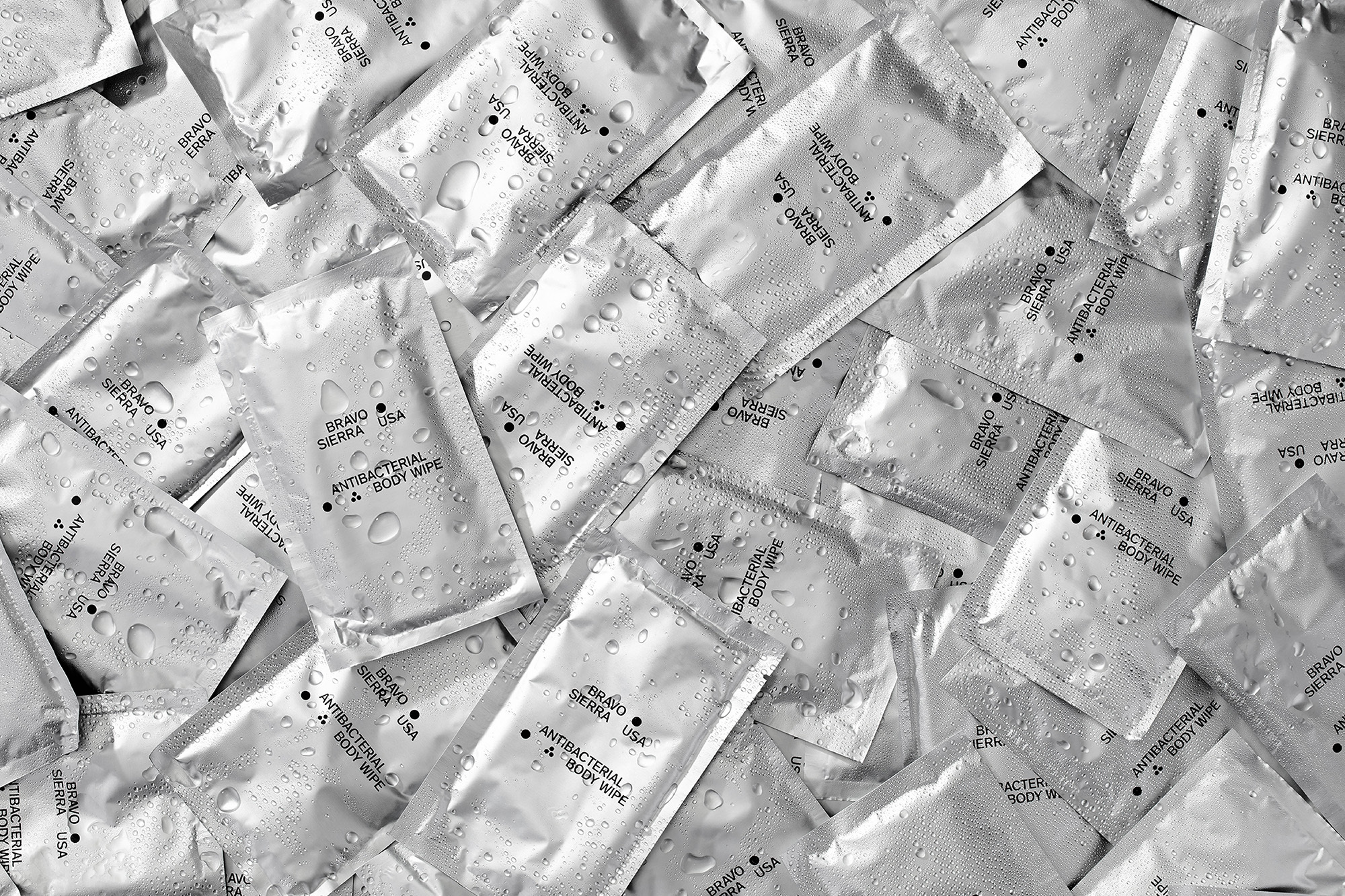 Close-up overhead shot of a silver foil packets of Bravo Sierra antibacterial body wipes with water droplets, filling the entire frame.