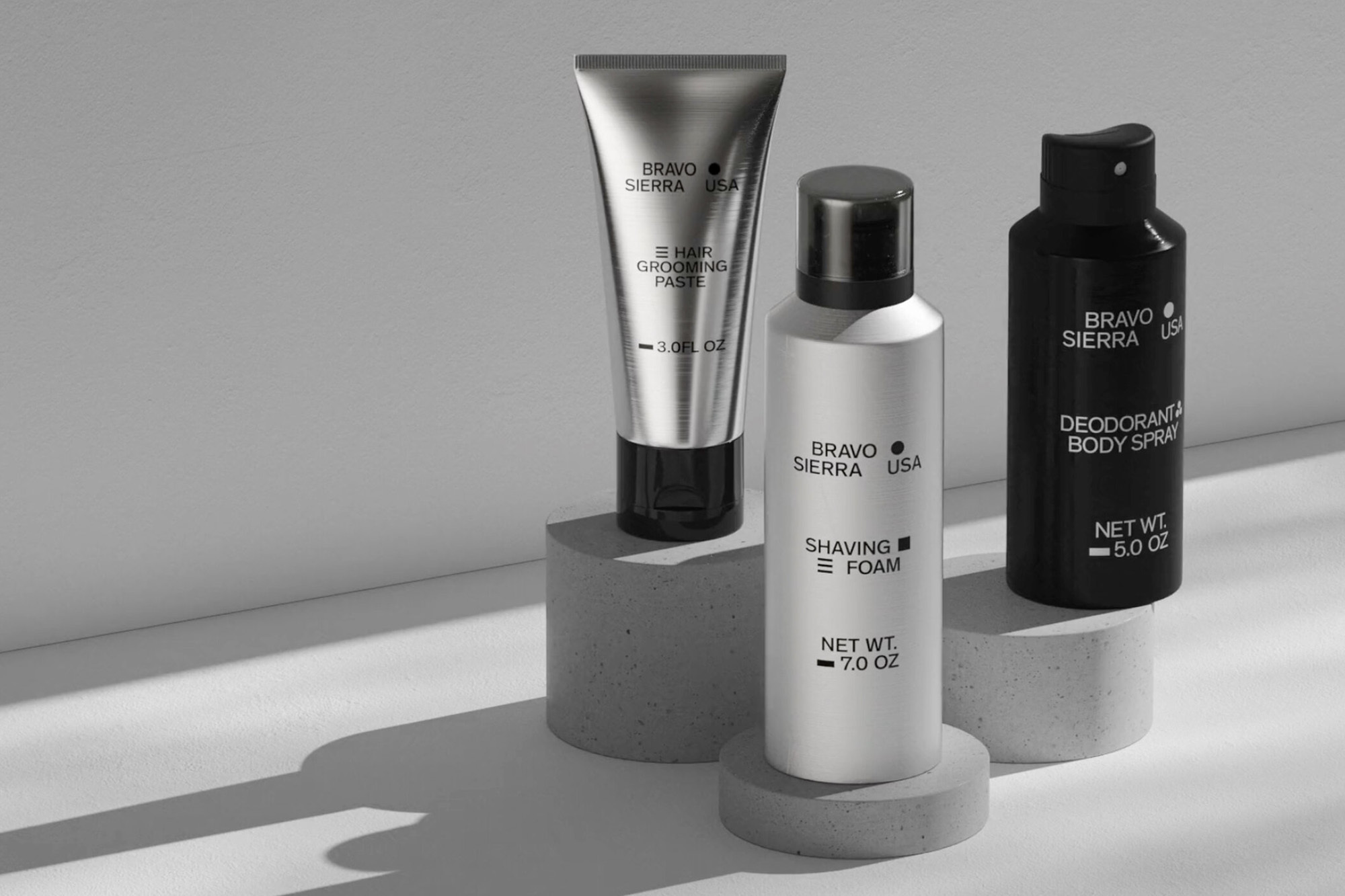 Silver tube of hair paste, white shaving foam bottle, and black deodorant spray on concrete pedestals in a high-key product shot.
