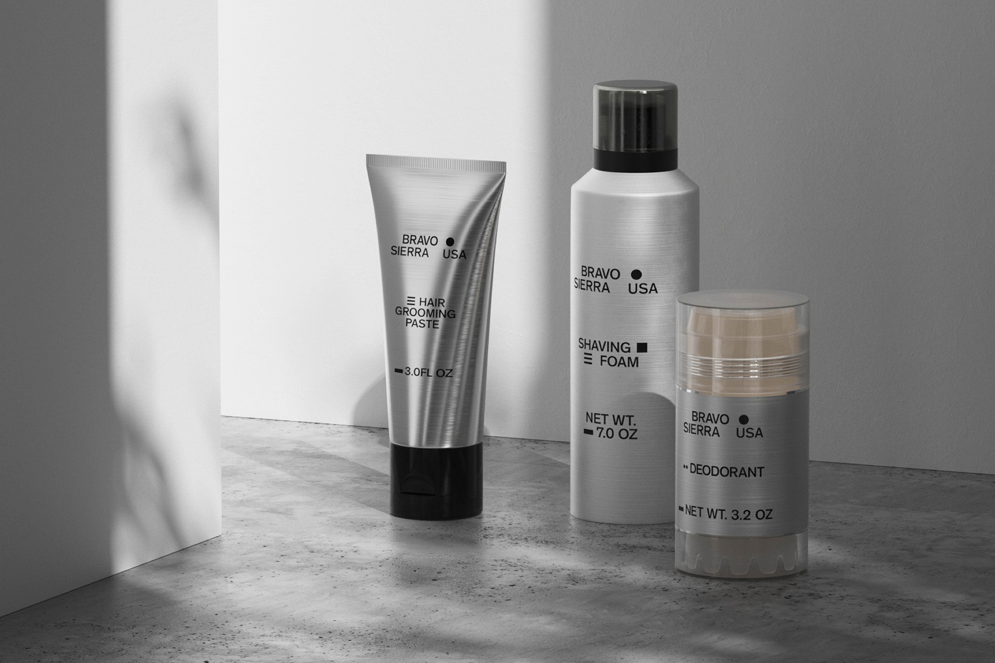 Bravo Sierra grooming products: hair paste, shaving foam, and deodorant on a gray surface against a white wall with shadows. 