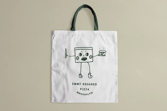 Canvas tote bag featuring cartoon monster graphic holding pizza burger with brand text, promoting Emmy Squared Pizza Brooklyn.