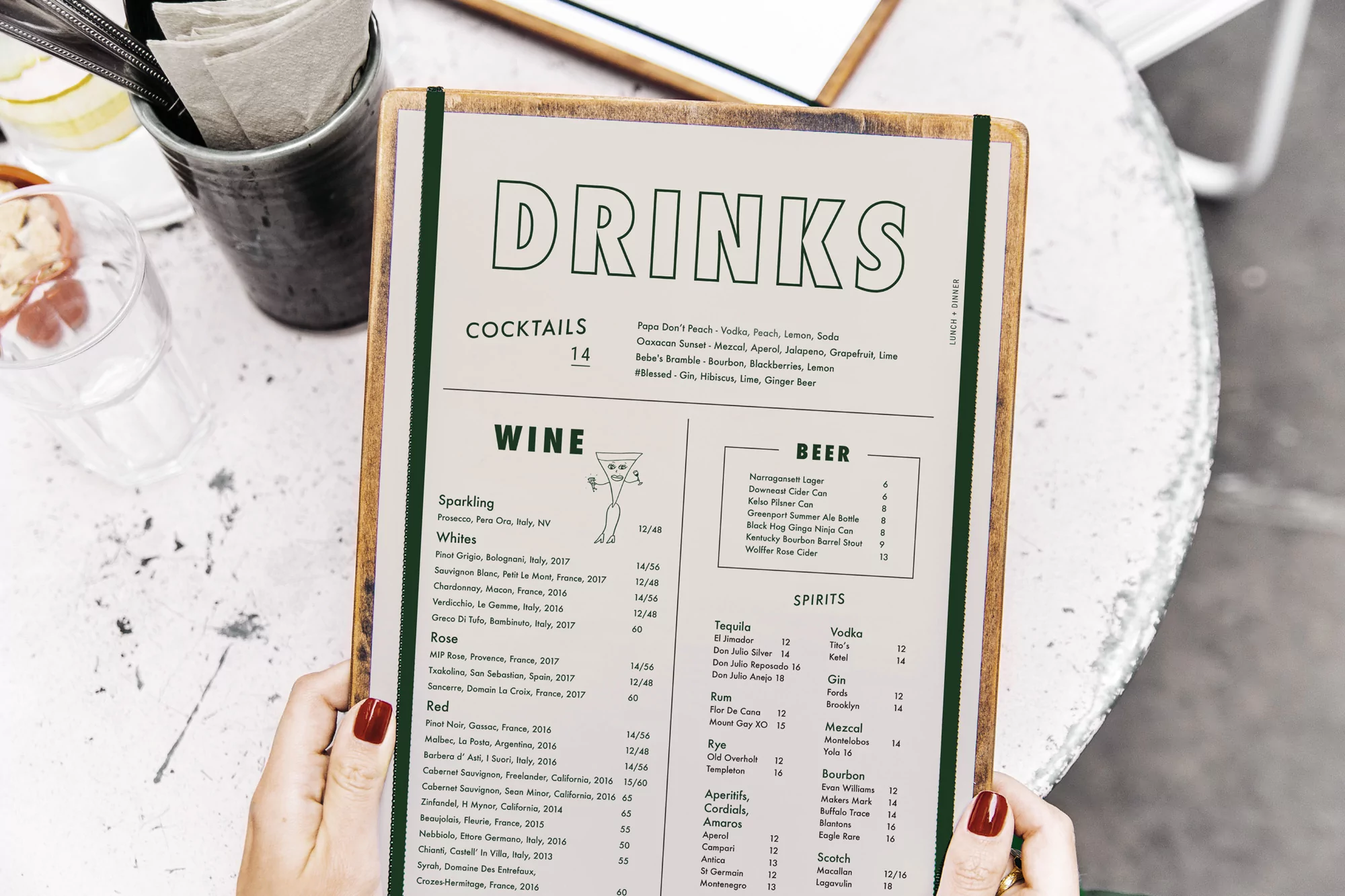 Close-up of a woman holding a drinks menu with visible cocktails, wine, beer, and spirits list on a marble-textured table in a cafe setting.