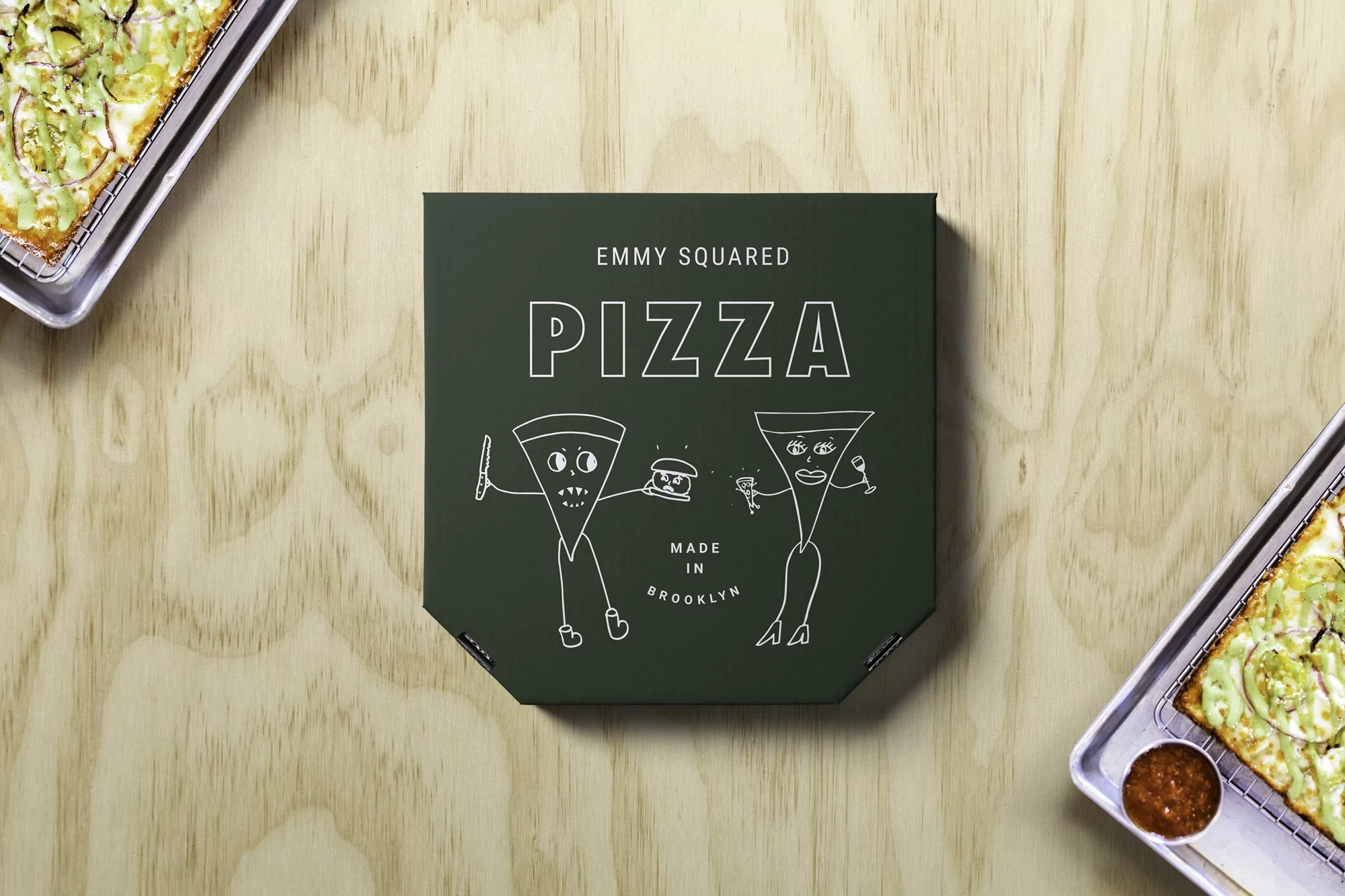 Overhead shot of a pizza box with cartoon pizza slices, resting on a light wood table with two trays of pizza on either side.