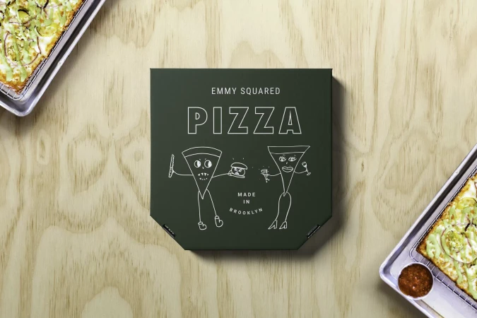 Overhead shot of a pizza box with cartoon pizza slices, resting on a light wood table with two trays of pizza on either side.