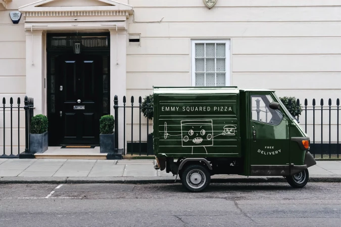 Dark green Emmy Squared Pizza delivery vehicle parked on a city street in front of a white building with a black door and window.