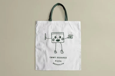 White canvas tote bag with dark green handles featuring a whimsical illustrated character holding a pizza slice and knife against a neutral background