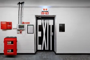 A black fire exit door with bold white EXIT typography set against a white concrete block wall with red safety equipment.