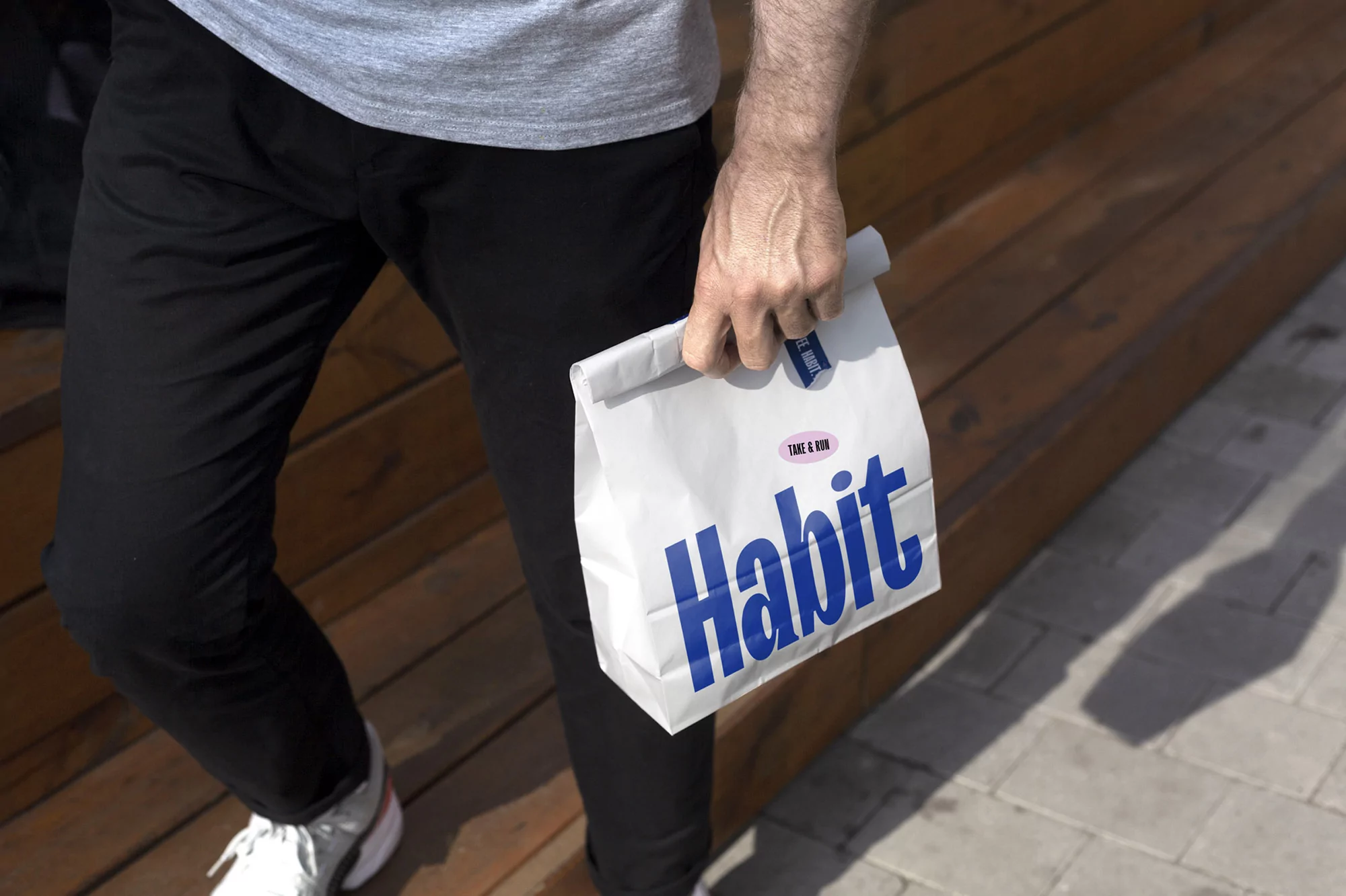 Adult male carries a white paper bag with a blue 'Habit' logo and text in an outdoor setting with wood and stone features.
