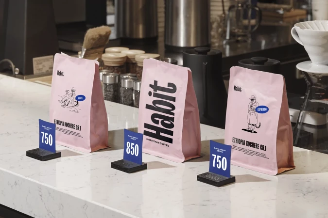 Three pink bags of Habit brand coffee beans are displayed on a counter with price tags in a bright, indoor coffee shop setting.