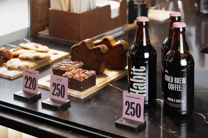 Assortment of baked goods on a counter with bottled cold brew coffee, including cookies, loaf cake, and brownies with nuts in a cafe.