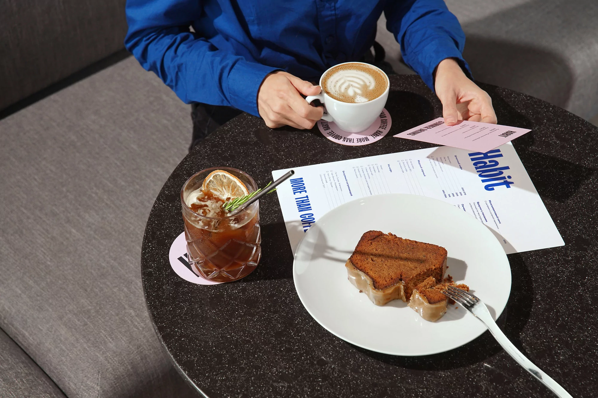 Person in a blue shirt sitting at a cafe table with coffee, cake, and a menu, enjoying a beverage and a pastry in a relaxed setting.