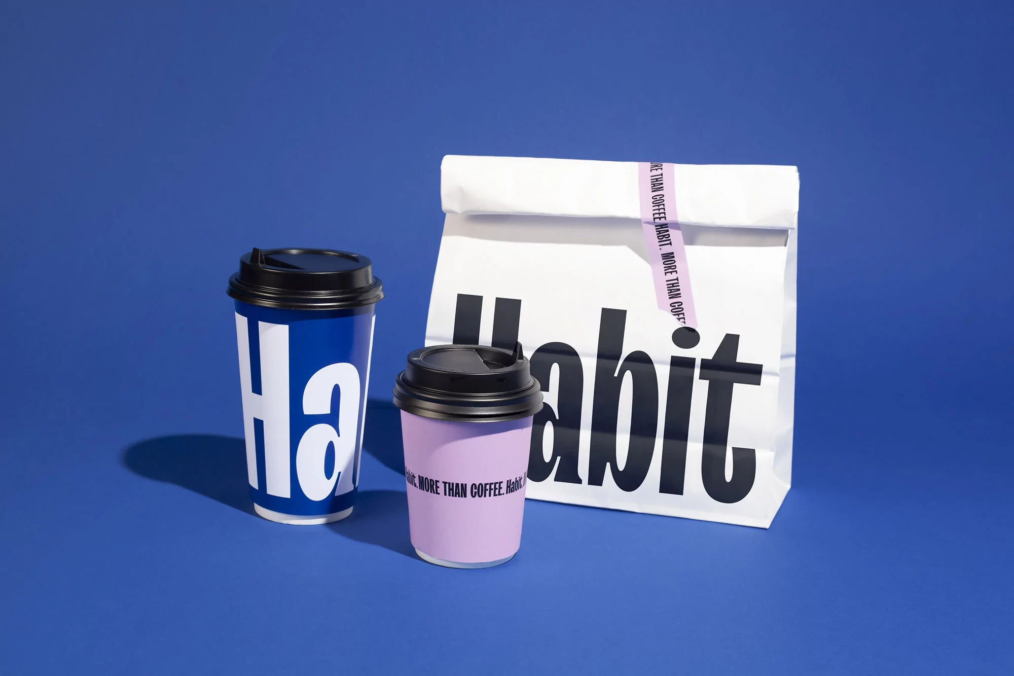 Two paper coffee cups and a white paper bag with logo on a blue background in a studio product shot for food and beverage.