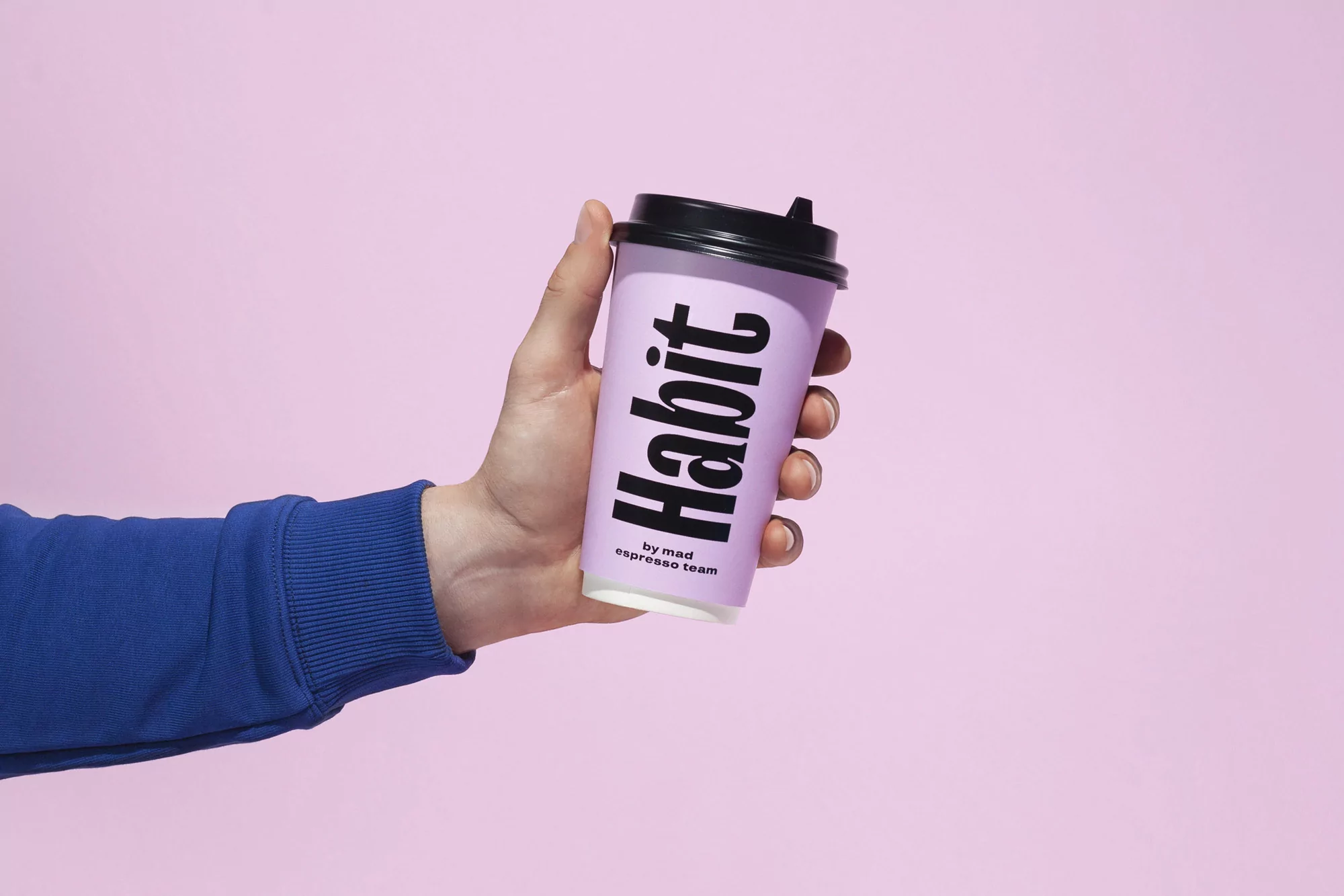 Close-up of a hand holding a lilac-colored disposable coffee cup with a black lid against a pink background, featuring the 'Habit' logo.