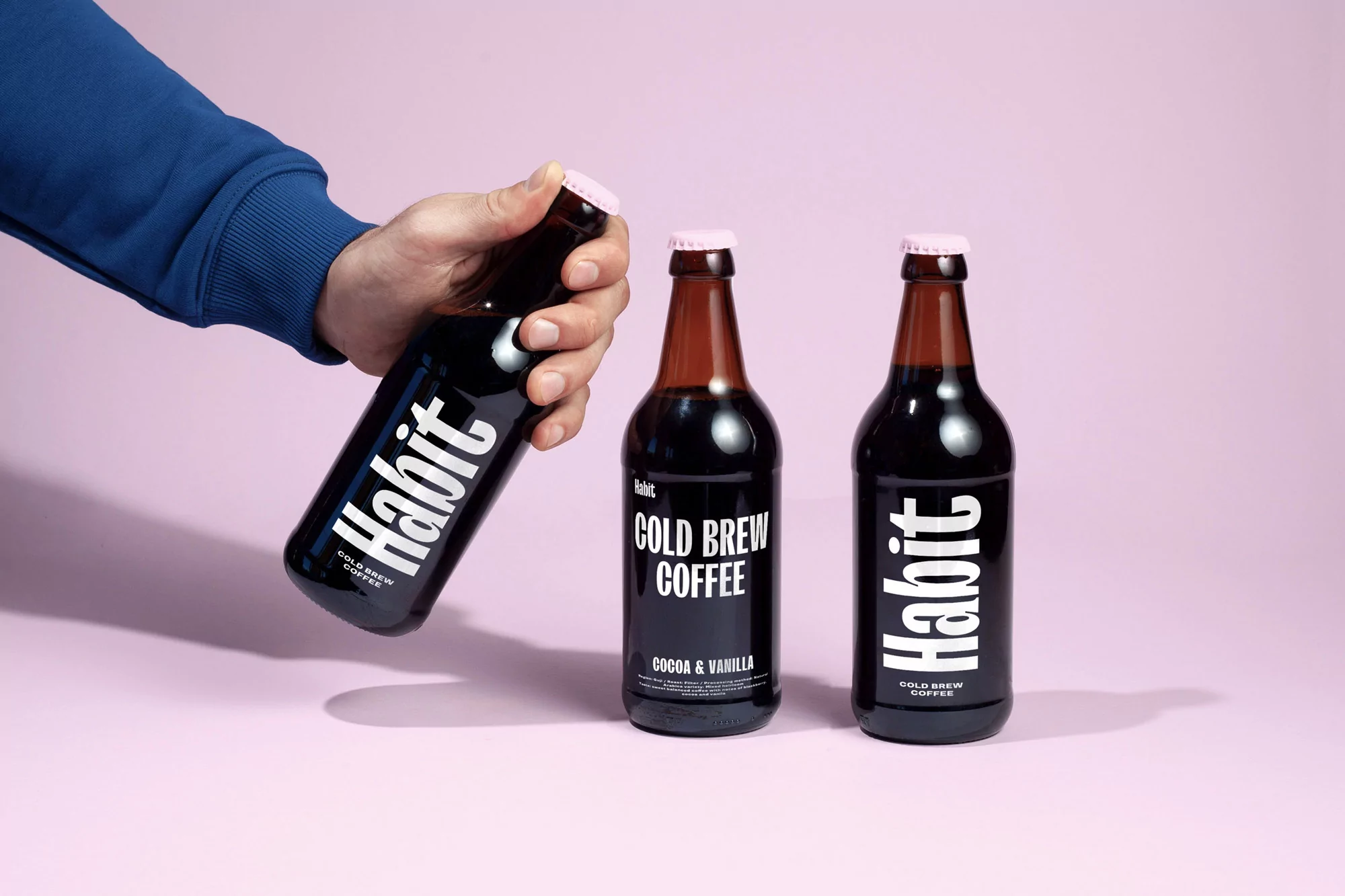 Hand holds a bottle of Habit cold brew coffee next to two other bottles, all against a pink background in a studio setting.