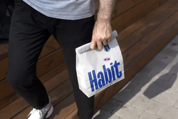 Man wearing grey t-shirt and black trousers holding a branded white paper bag from Habit