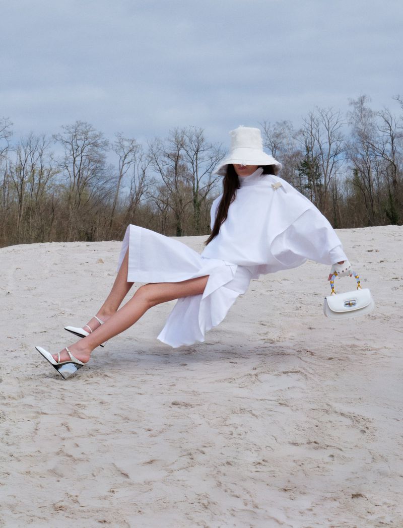 A woman in a white dress, hat, and high heels poses on a sandy surface with bare trees and a cloudy sky in the background.