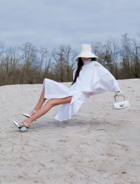 A woman in a white dress, hat, and high heels poses on a sandy surface with bare trees and a cloudy sky in the background.