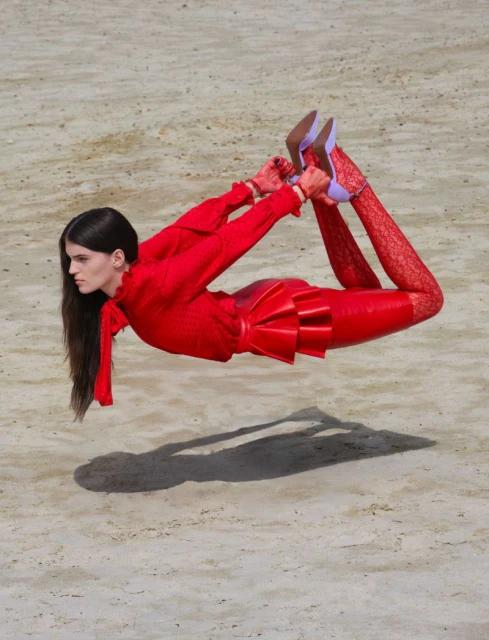 A woman in a red outfit performing a gymnastic pose, appearing to float above a sandy surface, creating an interesting visual.