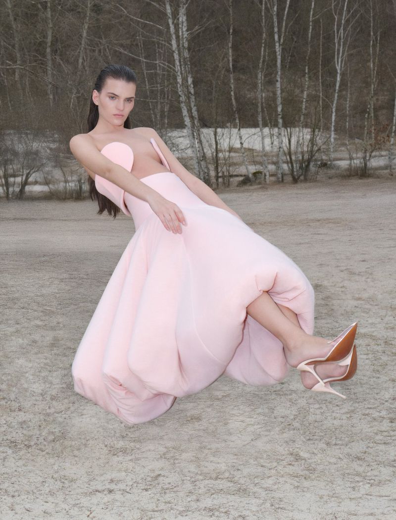 A young woman in a pink couture dress and heels strikes a pose against a blurry backdrop of bare trees and grey ground in a fashion shot.