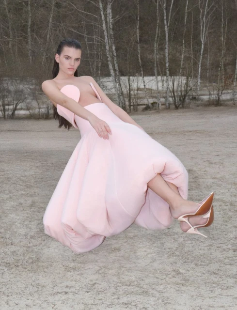 A young woman in a pink couture dress and heels strikes a pose against a blurry backdrop of bare trees and grey ground in a fashion shot.